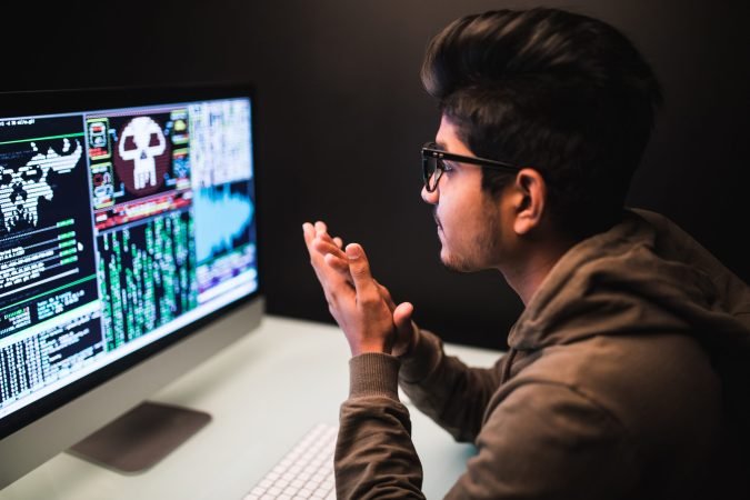 cybercrime, hacking and technology concept - male hacker with smartphone and coding on computers screen in dark room