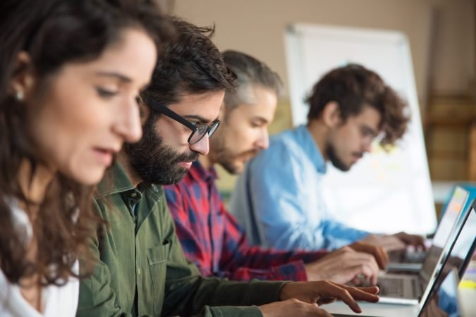 Line of coworkers using laptops in training room or class. Business colleagues in casual working together in contemporary office space. Workplace concept